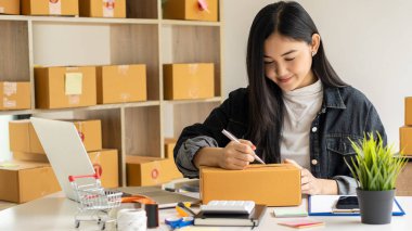Güzel Asyalı KOBİ işletme sahibi online alışverişte dizüstü bilgisayarda çalışıyor ve evdeki masada paket kutuları var - KME çevrimiçi iş fikri ve kutu teslimatı.