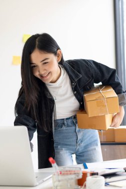Güzel Asyalı KOBİ işletme sahibi online alışverişte dizüstü bilgisayarda çalışıyor ve evdeki masada paket kutuları var - KME çevrimiçi iş fikri ve kutu teslimatı.