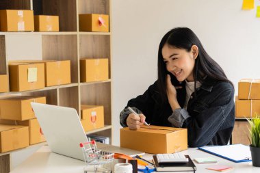 Asyalı bir kız iş kuruyor. Küçük işletme sahipleri iş yerinde dizüstü bilgisayarlar ve akıllı telefonlarla çalışıyorlar. Müşterilere çevrimiçi satış, e-ticaret, paketleme, ev işleri için paket karton kutular ile