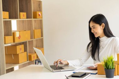 Asyalı bir kız iş kuruyor. Küçük işletme sahipleri iş yerinde dizüstü bilgisayarlar ve akıllı telefonlarla çalışıyorlar. Müşterilere çevrimiçi satış, e-ticaret, paketleme, ev işleri için paket karton kutular ile