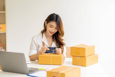Asyalı bir kız iş kuruyor. Küçük işletme sahipleri iş yerinde dizüstü bilgisayarlar ve akıllı telefonlarla çalışıyorlar. Müşterilere çevrimiçi satış, e-ticaret, paketleme, ev işleri için paket karton kutular ile