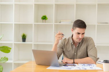 Genç işadamı, ofiste dizüstü bilgisayar ve hesap makinesiyle finansal rapor verilerini analiz ediyor ve beyin fırtınası yapıyor. Mali muhasebe ve yatırımla çalışma kavramı