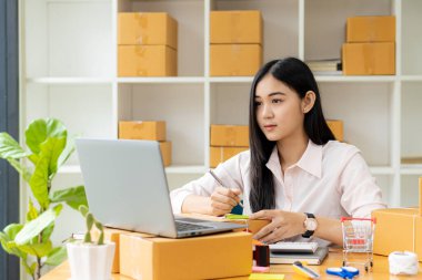 Küçük bir işletmenin portresi, KME Sahibi, Asyalı Kadın Girişimci Fatura kutuları üzerinde çalışır ve kutuları hazırlamak için çevrimiçi siparişleri işaretler. Müşterilere satıyorum. Çevrimiçi KOBİ iş fikri.