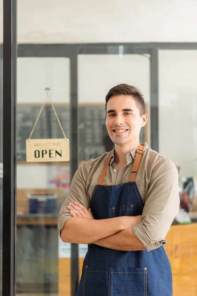 A handsome male waiter stood in front of his shop. Wearing a friendly apron, he smiles. beside the sign with the word open on the glass door