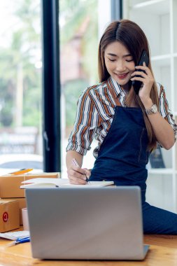 Asyalı kadın küçük bir KOBİ işine başlıyor. Kutular ve dizüstü bilgisayarlar üzerinde çalışıyor. Nakliye için paketler hazırlıyor. Evden çalışma, paket toplama, kutuları dikey sırayla paketleme kavramı.