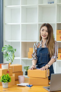 Asyalı kadın küçük bir KOBİ işine başlıyor. Kutular ve dizüstü bilgisayarlar üzerinde çalışıyor. Nakliye için paketler hazırlıyor. Evden çalışma, paket toplama, kutuları dikey sırayla paketleme kavramı.