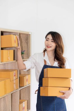 Asyalı kadın küçük bir KOBİ işine başlıyor. Kutular ve dizüstü bilgisayarlar üzerinde çalışıyor. Nakliye için paketler hazırlıyor. Evden çalışma, paket toplama, kutuları dikey sırayla paketleme kavramı.