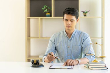 Asyalı erkek avukat bir mahkeme salonunda yasal sözleşme belgeleri üzerinde çalışıyor. Masanın üzerinde çekiç ve teraziyle hukuk kavramları.