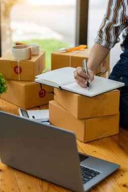 Evde kutu ve dizüstü bilgisayarla çalışan genç bir kadın çevrimiçi iş konsepti paketleri teslim etmeye hazır müşterilerden emir alıyor.