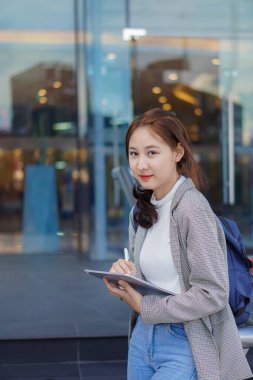 Bir alışveriş merkezinin önünde elinde tablet ve çantasını tutan güzel, gülümseyen bir kız öğrenci..