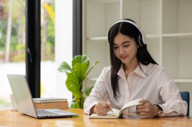 Bilgisayarlı dijital kurslar geliştirecek programları dinlemek için kulaklık takan güzel bir Asyalı kadın. Evdeki sosyal mesafeler, kariyer yolunda sınıfta beceriler geliştirir.
