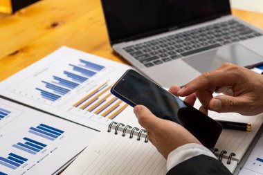 Akıllı telefon ve bilgisayarla çalışan işadamı, kazancı hesaplamak için finansal verileri ve hesap makinesini analiz etmek için bilgisayarla çalışıyor..