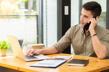 Genç işadamı ofisteki masadaki mali grafikleri analiz etmek için akıllı telefon ve laptop kullanıyor.