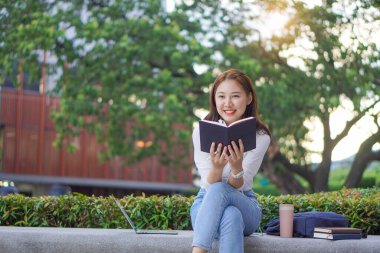 Bir kız üniversite öğrencisi kaplan derisi ve yanında kaplan derisi bir laptop çantası olan bir kupayı tutarken bulanık zeminde mutlu bir şekilde gülümsüyor. Açık hava eğitim değişim öğrencisi konsepti