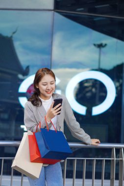 Neşeli, gülümseyen Asyalı genç bir kadın alışveriş merkezinin önünde dururken çok renkli bir alışveriş çantası ve cep telefonu tutuyor. Yaşam tarzı teknoloji kavramı