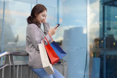Neşeli, gülümseyen Asyalı genç bir kadın alışveriş merkezinin önünde dururken çok renkli bir alışveriş çantası ve cep telefonu tutuyor. Yaşam tarzı teknoloji kavramı