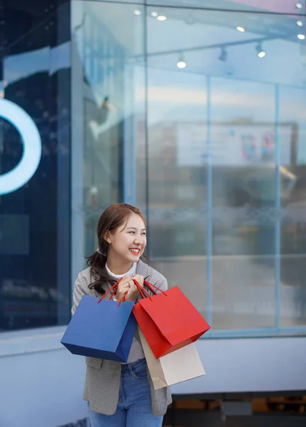 明るく笑顔の10代のアジアの女性は ショッピングモールの前に立つと 多色のショッピングバッグと携帯電話を持っています ライフスタイル技術の概念 — ストック写真