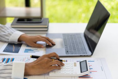 Asyalı bir muhasebecinin eli hesap makinesine basıyor ve hemen yanındaki mali grafikleri ve bilgisayarı analiz ederken aynı zamanda elinde bir kalem tutuyor..