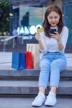 Neşeli gülümseyen güzel Asyalı kadın elinde bir kredi kartı tutuyor ve alışveriş merkezinin arka planında alışveriş torbalarıyla online alışveriş yapmak için akıllı telefon kullanıyor..