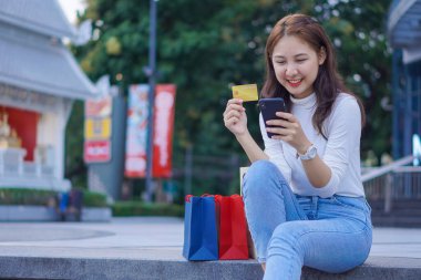 Neşeli gülümseyen güzel Asyalı kadın elinde bir kredi kartı tutuyor ve alışveriş merkezinin arka planında alışveriş torbalarıyla online alışveriş yapmak için akıllı telefon kullanıyor..