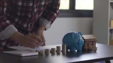 Para biriktirmek, ev almak, finansal ve ekonomik kavramlar için kumbarada para sayan genç bir adam. Evdeki hesap makineleri ve vergi belgeleri kullanılarak aylık harcamalar ve borç geri ödemeleri.
