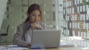 Beyaz odada cep telefonu kullanan Asyalı bir kadın cep telefonu kullanıyor çevrimiçi ödemeler ve ofiste bilgisayar ve grafik belgeleriyle birlikte iş konsepti tablolarının hesaplama analizinde..