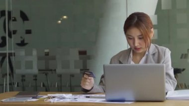 Beyaz odada cep telefonu kullanan Asyalı bir kadın cep telefonu kullanıyor çevrimiçi ödemeler ve ofiste bilgisayar ve grafik belgeleriyle birlikte iş konsepti tablolarının hesaplama analizinde..