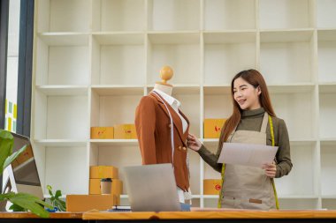 Gülümseyen Asyalı kadın moda tasarımcısı oldukça iyi görünümlü bir atölyede dizüstü bilgisayarda çalışıyor, çalışıyor ve internette el yapımı KME iş fikirlerinin şık koleksiyonunu inceliyor..