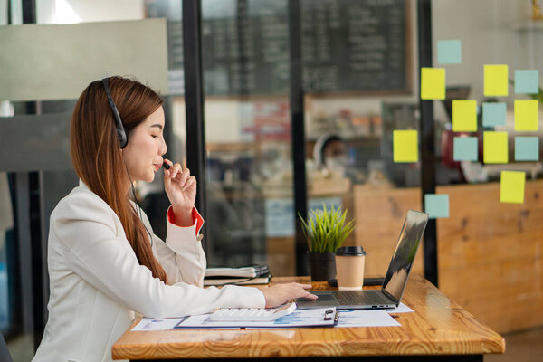 Asian woman wearing headphones using laptop talking to a coworker about sales report in video conferencing while working at a coffee shop. financial accounting business concept