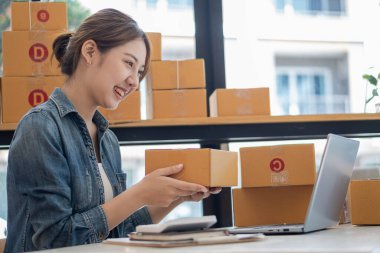 Küçük bir işletmenin portresi, KOBİ sahibi, bayan girişimci Asyalı kız kutular ve dizüstü bilgisayarlar üzerinde çalışıyor, müşterilere satılacak kutular için çevrimiçi siparişleri kontrol ediyor. Çevrimiçi KOBİ iş fikri.