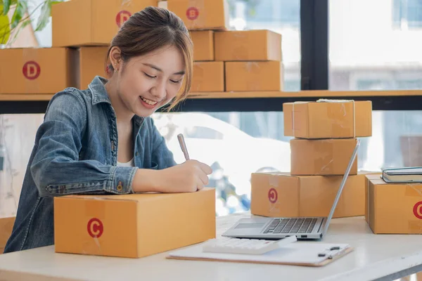 Küçük bir işletmenin portresi, KOBİ sahibi, bayan girişimci Asyalı kız kutular ve dizüstü bilgisayarlar üzerinde çalışıyor, müşterilere satılacak kutular için çevrimiçi siparişleri kontrol ediyor. Çevrimiçi KOBİ iş fikri.