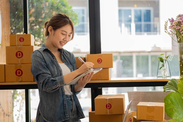 Küçük bir işletmenin portresi, KOBİ sahibi, bayan girişimci Asyalı kız kutular ve dizüstü bilgisayarlar üzerinde çalışıyor, müşterilere satılacak kutular için çevrimiçi siparişleri kontrol ediyor. Çevrimiçi KOBİ iş fikri.