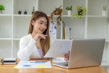 Güzel bir Asyalı kadın elinde grafik dokümanlarıyla ofiste dizüstü bilgisayarla çalışıyor. Finansal istatistik veri analizi grafiği büyüyen iş dünyası fikrine bakan iş kadını