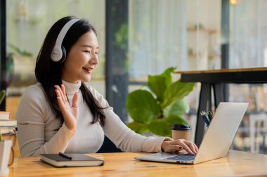 Asya kökenli, kulaklıkla dizüstü bilgisayara bakmaktan mutlu bir bayan yönetici. İnternette müşterilerle mutlu bir şekilde sohbet et. İşyeri eğitim konseptinde uzaktan çalışmak bir kahve dükkanında çalışmak