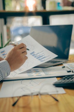 Masada oturan bir işadamı elinde kalemle finans tablosunu analiz ediyor. Muhasebeciler iş raporlarında hesaplanan veri tablolarıyla çalışıyorlar. Ofiste bir dizüstü bilgisayarla.