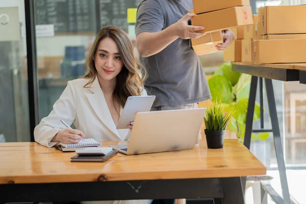 Gülümseyen Asyalı kadın kutulara bakar ve onları müşterilere göndermeden önce paket teslimat kutularındaki bilgileri kontrol etmek için dizüstü bilgisayar kullanır. Girişimciler ve küçük işletmeler çevrimiçi pazarlamada çalışıyor