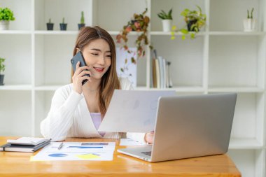 Dizüstü bilgisayarla tasarruf, finans ve ekonomi kavramlarını hesaplayan Asyalı kadın muhasebeci ya da bankacı. Bir iş kadını akıllı telefon kullanır ve sonuçları gösteren çizelgeler ve grafikler yazar..
