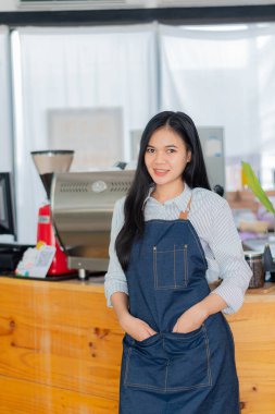 Önlüklü genç bir Asyalı barista bir işletme sahibinin başlangıç konseptinin önünde duruyor. KOBİ kafe kasiyerini yöneten başarılı kadın.