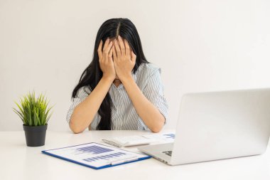 Mavi gömlekli genç bir Asyalı iş kadını elleriyle yüzünü örtüyor ve finansal muhasebe ofisinde dizüstü bilgisayar ve grafik kağıdıyla çalışırken strese giriyor..