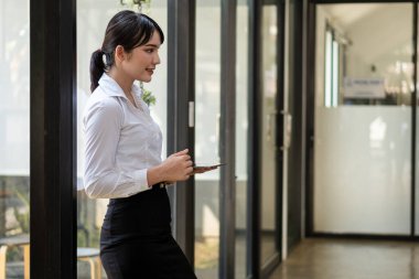 Ofiste büyük cam pencereli bir yerde duran Asyalı bir kadın, işini düzenlemek için dijital ped kullanıyor..