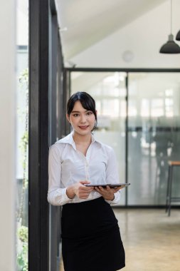 Ofiste büyük cam pencereli bir yerde duran Asyalı bir kadın, işini düzenlemek için dijital ped kullanıyor..