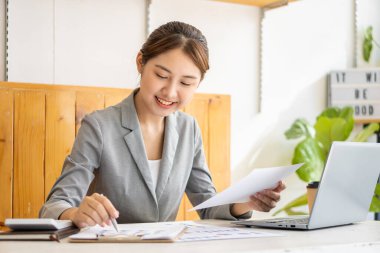 Güzel Asyalı iş kadını hesap makinesi kullanarak analiz çizelgesi, ofiste elinde kağıtlar ve elinde kalemle modern ev stüdyosundaki sandalyede oturuyor. Cep telefonu işçisi müşterilerle bağlantı kuruyor