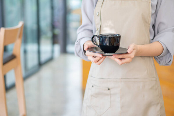 A young woman barista holding a cup of coffee serving customers at a coffee shop employee offers hot coffee in a modern shop.