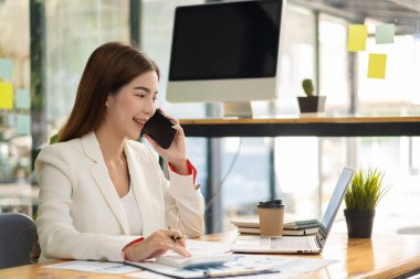 Asyalı iş kadını, masada duran çalışma grafiği belgesini hesaplamak için istatistiksel analizi hesaplayan bir hesap makinesiyle masada iş görüşmesi yapmak için akıllı telefon kullanıyor.