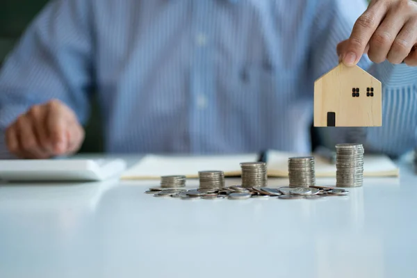 A man holds a house with piles of coins on the table saving money to spend and arrange a future home purchase. Real Estate Investment Ideas and Low-Interest Rate Home Loans for Homes