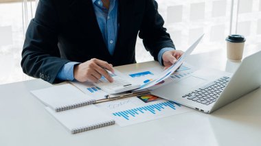 İş adamı ya da muhasebeci elinde kalem ve analitik çizelge tutan kağıtlar şirket bütçesini ve mali planlama ciro oranını grafik raporuyla hesaplar ve veri analizi için dizüstü bilgisayarı kullanır.