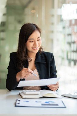 Kağıt üzerinde çalışan, elinde kalem ve laptop tutan gülümseyen Asyalı iş kadını modern ev stüdyosunda bir sandalyede oturuyor. İş yerindeki mobil işçiler ve hesap makineleri Finans veya Muhasebe kavramları