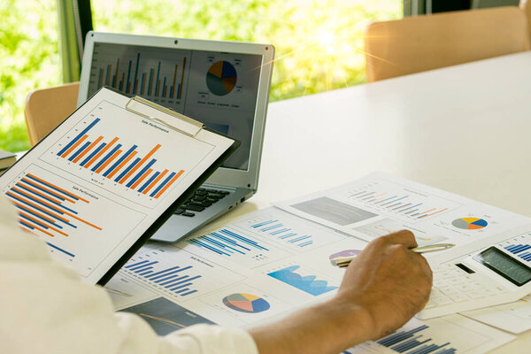 Бизнесмен с ручкой, указывающий на график, финансовые данные, исследования, разработки, отчет по рынку. Стратегическое планирование, анализ и расчет финансовых счетов с помощью калькулятора и ноутбука в офисе.