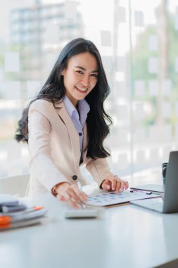 Ofisteki mali hesabı analiz etmek için hesap makinesi kullanan Asyalı iş kadını, finansal grafik belgesi üzerinde çalışıyor.