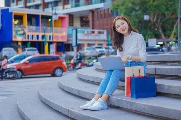 亚洲女人 提着五颜六色的购物袋 开心地微笑着 坐在购物中心前面 — 图库照片